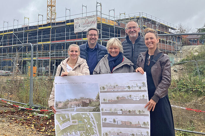 Fünf Personen halten den Bauplan für den Anbau der Regenbogenschule Stolberg hoch. Im Hintergrund ist die Baustelle.