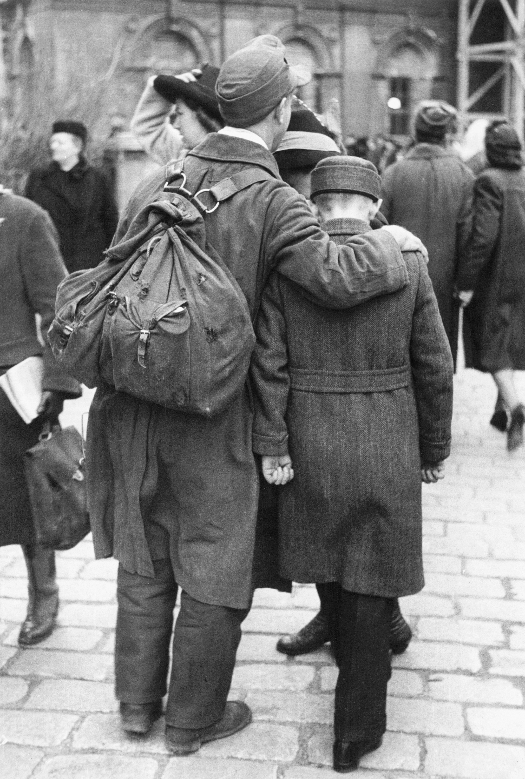 Aufnahme in Schwarz-Weiß, die zwei Männer von hinten zeigt. Der linke trägt einen Rücksack und umarmt den kleineren Jungen rechts.